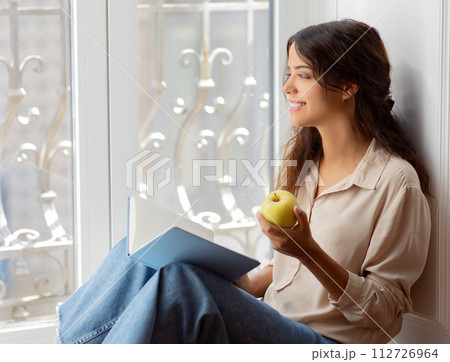 Smiling young woman enjoying healthy snack and reading book by the window 112726964