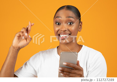 Hopeful black woman poses crossing fingers while using smartphone, studio 112727289