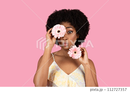 Playful young woman with curly hair making a funny face, holding a pink gerbera over her eye Playful young woman with curly hair making a funny face, holding a pink gerbera over her eye 112727357