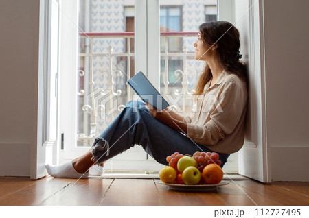 Serene young woman reading book beside bright window at home Serene young woman reading book beside bright window at home 112727495