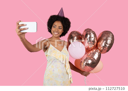 Vibrant African American woman with curly hair and a party hat taking a selfie 112727510