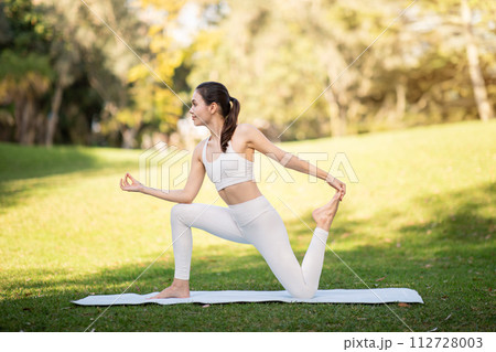 A focused young woman in a white yoga outfit holds an elegant dancer's pose on a mat in a sunny 112728003