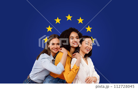 Three joyful women of diverse ethnicities hugging and smiling in front of a European Union flag 112728045