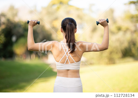 Rear view of a fit woman exercising with dumbbells in a sunny park, showcasing her strength and healthy lifestyle Rear view of a fit woman exercising with dumbbells in a sunny park, showcasing her strength and healthy lifestyle 112728134