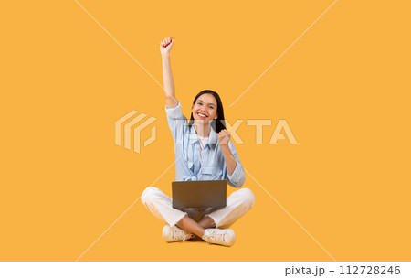 Joyful woman with laptop celebrating success on yellow backdrop 112728246