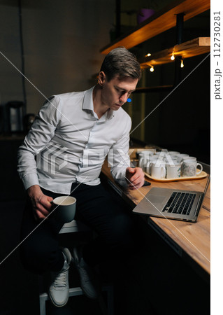 Vertical portrait of focused male reading on laptop working on deadline during late shift. Business, online and corporate worker drinking coffee and doing research on computer during overtime at work. 112730281