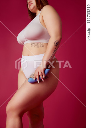 Cropped portrait of young overweight woman in inner wear holds in hands blue toy for adults against vibrant pink studio background. 112735219