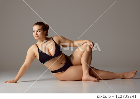 Smiling woman with visible tattoo on body reclining wearing dark bikini and holding hand on knee against grey studio background. Smiling woman with visible tattoo on body reclining wearing dark bikini and holding hand on knee against grey studio background. 112735241