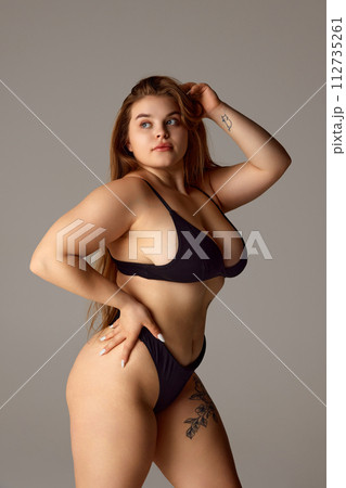 Portrait of beautiful, overweight woman with in dark lingerie posing confidently holds hand on hip against grey studio background. 112735261