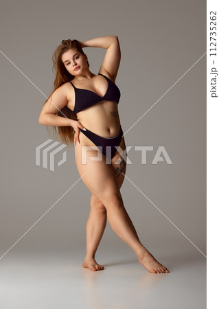 Full-length portrait of woman with tattoo on thigh in dark lingerie posing confidently holds hand on hip against grey studio background. Full-length portrait of woman with tattoo on thigh in dark lingerie posing confidently holds hand on hip against grey studio background. 112735262