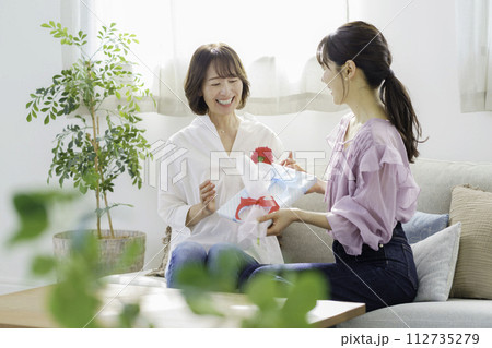 母の日　自宅のリビングででカーネーションとプレゼントを渡す若い女性と受け取る母親 112735279
