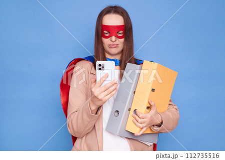 Unhappy woman wearing superhero costume standing isolated over blue background holding folders with documents using mobile phone reading bad news browsing Unhappy woman wearing superhero costume standing isolated over blue background holding folders with documents using mobile phone reading bad news browsing 112735516