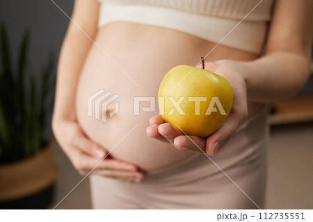 Unknown beautiful pregnant woman with bare belly standing in home interior holding fresh organic yellow apple prefers healthy eating while expecting baby Unknown beautiful pregnant woman with bare belly standing in home interior holding fresh organic yellow apple prefers healthy eating while expecting baby 112735551