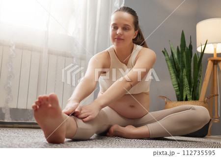 Caucasian pregnant woman wearing beige top and leggings exercising at home stretching her legs enjoying gymnastic exercising in light living room interior Caucasian pregnant woman wearing beige top and leggings exercising at home stretching her legs enjoying gymnastic exercising in light living room interior 112735595