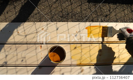 hand with a brush and a jar of brown paint on a wooden surface of boards in bright sunlight, top view, still life with objects for painting and processing wooden building material 112736407