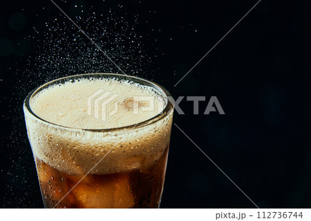 Glass wit foamy drink, soda water, coke, dark beer with ice cubes against black background. Glass wit foamy drink, soda water, coke, dark beer with ice cubes against black background. 112736744