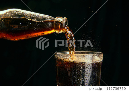 Soda, coke, dark beer pouring from bottle into glass against black background 112736750