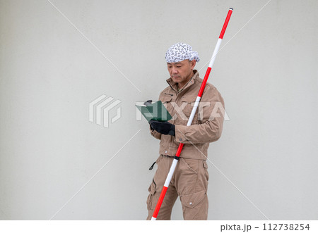 測量ポールを小脇に抱えながらメモ帳に記入をしている土木系の男性 測量ポールを小脇に抱えながらメモ帳に記入をしている土木系の男性 112738254