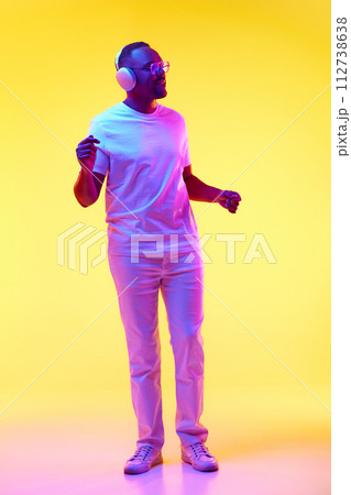 Full-length of African-American man in casual clothes listening to music in headphones and dancing against yellow studio background in neon light Full-length of African-American man in casual clothes listening to music in headphones and dancing against yellow studio background in neon light 112738638
