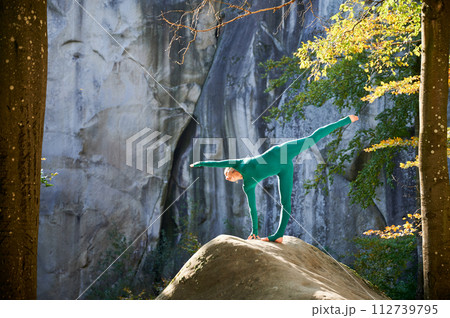 Woman practicing yoga outdoors in forest. Barefoot female on yoga mat surrounded by trees and large rocks, which suggests peaceful, natural environment ideal for meditation or yoga practice. 112739795
