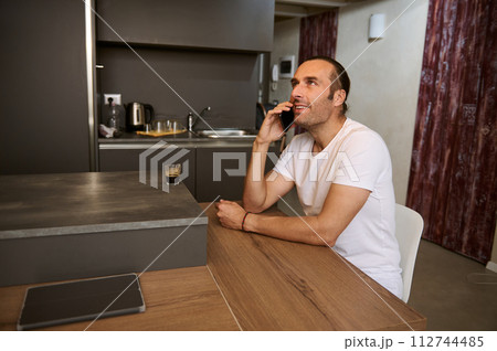 Smiling man talking on mobile phone, sitting at table at home kitchen Smiling man talking on mobile phone, sitting at table at home kitchen 112744485