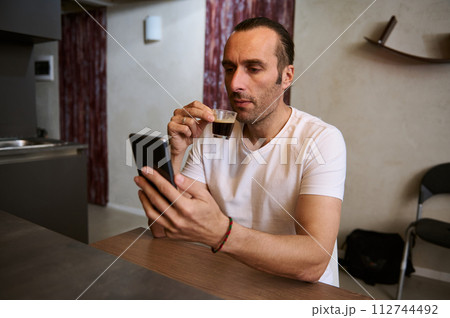 Authentic portrait of young Caucasian handsome man drinks coffee and reads news, sitting at table in the home interior Authentic portrait of young Caucasian handsome man drinks coffee and reads news, sitting at table in the home interior 112744492