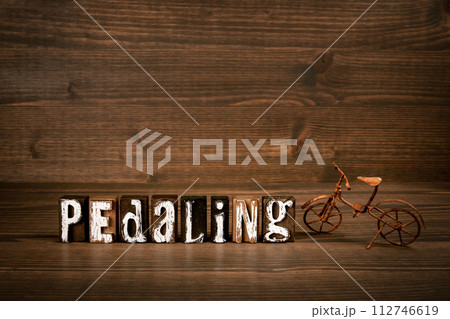 PEDALING. Text from alphabet blocks and rusty miniature bicycle on wood texture background 112746619