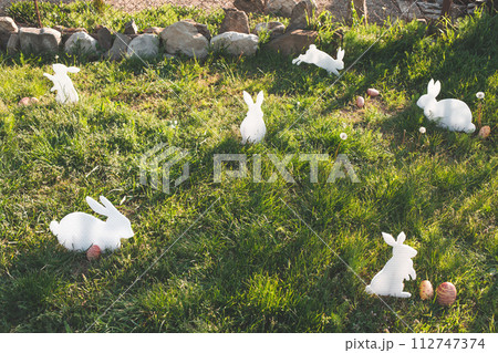 The concept of Easter holidays. Decorative handmade crafts in the form of white rabbits and colorful eggs. Grass in the background. Copy space The concept of Easter holidays. Decorative handmade crafts in the form of white rabbits and colorful eggs. Grass in the background. Copy space 112747374