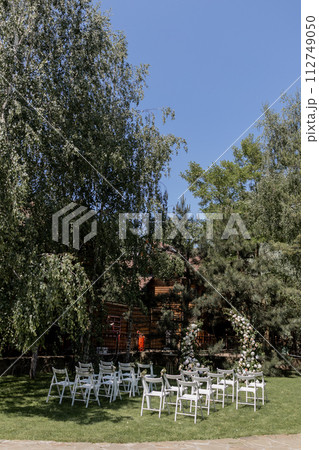 Outdoor wedding ceremony with a stunning floral arch and white chairs set in a serene garden 112749050