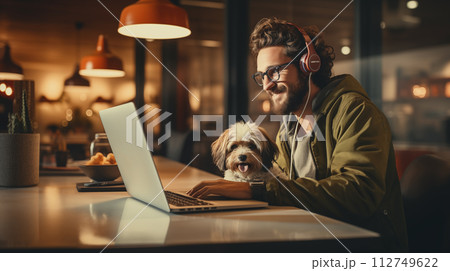 AI-generated content. A stylish man with a beard and headphones is sitting at a table with a laptop and a dog, in the evening light 112749622