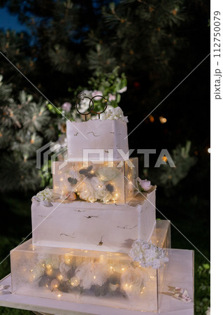 Elegant tiered wedding cake illuminated with romantic lights, set against a floral outdoor backdrop Elegant tiered wedding cake illuminated with romantic lights, set against a floral outdoor backdrop 112750079