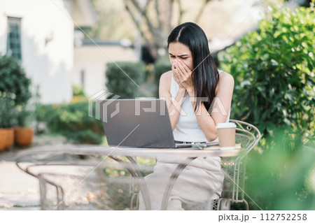 Worried young woman covering her mouth, looking at her laptop screen with concern, seated at a garden table with a coffee cup. Worried young woman covering her mouth, looking at her laptop screen with concern, seated at a garden table with a coffee cup. 112752258