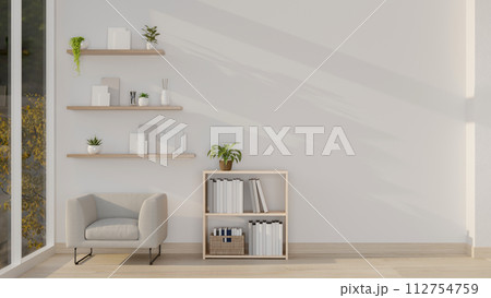 A minimalist apartment living room features a comfortable armchair, a bookcase, and a white wall 112754759