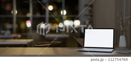 A table features a white-screen laptop computer mockup in a modern office at night. A table features a white-screen laptop computer mockup in a modern office at night. 112754769