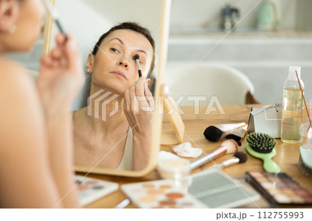 Middle-aged woman using brush during eye makeup sitting in front of the mirror 112755993