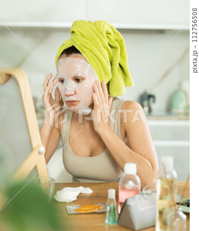 Middle-aged woman applying tissue mask on her facial skin sitting in front of the mirror 112756508
