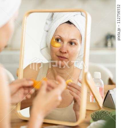 Woman in towel turban applying golden collagen eye patches Woman in towel turban applying golden collagen eye patches 112756552