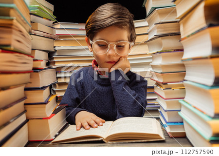 Cute little pupil boy in glasses reading interesting book in library between stacks of books Cute little pupil boy in glasses reading interesting book in library between stacks of books 112757590