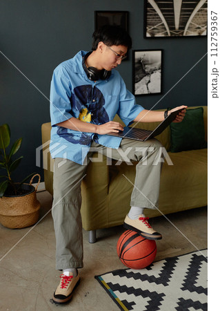 Vertical full length portrait of Asian young man using laptop at home or in office lounge and playing with basketball 112759367