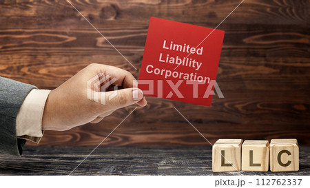 Business hand holding a note with LLC text, the acronym of Limited Liability Company with a row of wooden cubes with LLC text 112762337