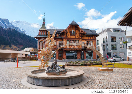 A serene Engelberg scene with a fountain sculpture, a Swissstyle building with a gabled roof, and contrasting modern architecture, set against snowcapped mountains and a clear sky. A serene Engelberg scene with a fountain sculpture, a Swissstyle building with a gabled roof, and contrasting modern architecture, set against snowcapped mountains and a clear sky. 112765443