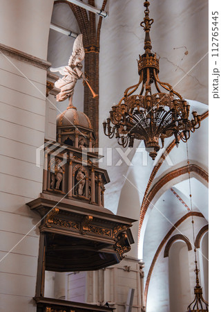 An ornate Riga Dome Cathedral interior with a gold accented wooden pulpit, white angel statue, and a large bronze chandelier, set against white walls and a red brick vaulted ceiling. 112765445
