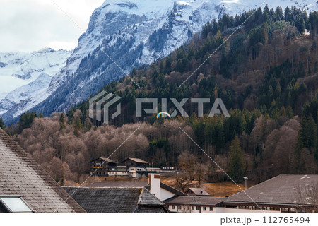 Scenic view of Engelberg, Switzerland, featuring a building with grey shingles, a dense coniferous forest, and snowcapped mountains under an overcast sky, with a colorful paraglider above. 112765494