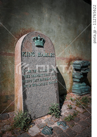 An aged stone memorial marked KING HAMLET with a mysterious epitaph and crown symbol, rests by a wall near old green books, likely in Helsingborg, Denmark. An aged stone memorial marked KING HAMLET with a mysterious epitaph and crown symbol, rests by a wall near old green books, likely in Helsingborg, Denmark. 112765622