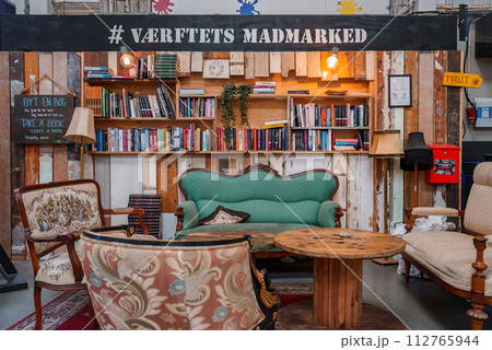 A cozy, eclectic indoor seating area with a green sofa, floral armchair, and rustic table, surrounded by bookshelves and wooden decor, likely in a Helsingborg food market. A cozy, eclectic indoor seating area with a green sofa, floral armchair, and rustic table, surrounded by bookshelves and wooden decor, likely in a Helsingborg food market. 112765944