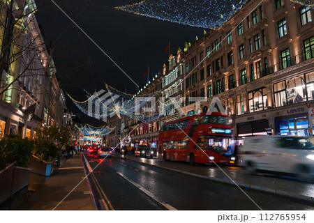 A bustling London Regent street at Christmas time, adorned with festive lights that mimic a starry sky. The scene includes iconic red buses, lively shops, and a warm, celebratory atmosphere. 112765954