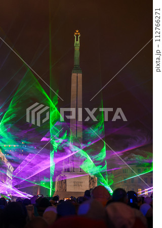 Nighttime celebration at Riga's Freedom Monument, Latvia, with green laser lights creating a festive atmosphere among a crowd of spectators dressed for cool weather. 112766271