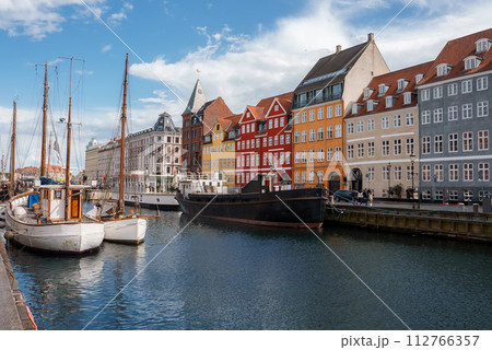 A serene day at Nyhavn, Copenhagen, with boats moored in the canal, reflecting the colorful facades of European townhouses, under a blue sky with scattered clouds. A serene day at Nyhavn, Copenhagen, with boats moored in the canal, reflecting the colorful facades of European townhouses, under a blue sky with scattered clouds. 112766357