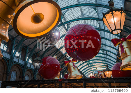 Inside a historic London venue, Covent Garden Market, oversized red baubles with golden bows and bellshaped lights create a warm, festive atmosphere under a curved glass roof. 112766490