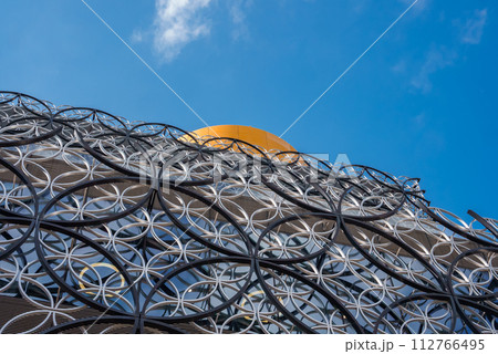 Closeup of the Library of Birmingham's metallic facade with interlocking circles and a bright yellow sphere against a clear blue sky, showcasing its modern design. 112766495